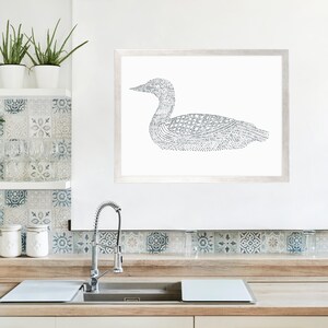 Puede incluir: Una impresi&oacute;n en blanco y negro de un p&aacute;jaro loon en arte de l&iacute;nea en un marco blanco. La impresi&oacute;n est&aacute; colgada en una pared blanca sobre una encimera de cocina con un fregadero y un salpicadero de azulejos.