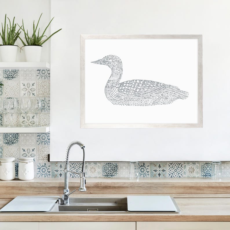 Puede incluir: Una impresi&oacute;n en blanco y negro de un p&aacute;jaro loon en arte de l&iacute;nea en un marco blanco. La impresi&oacute;n est&aacute; colgada en una pared blanca sobre una encimera de cocina con un fregadero y un salpicadero de azulejos.