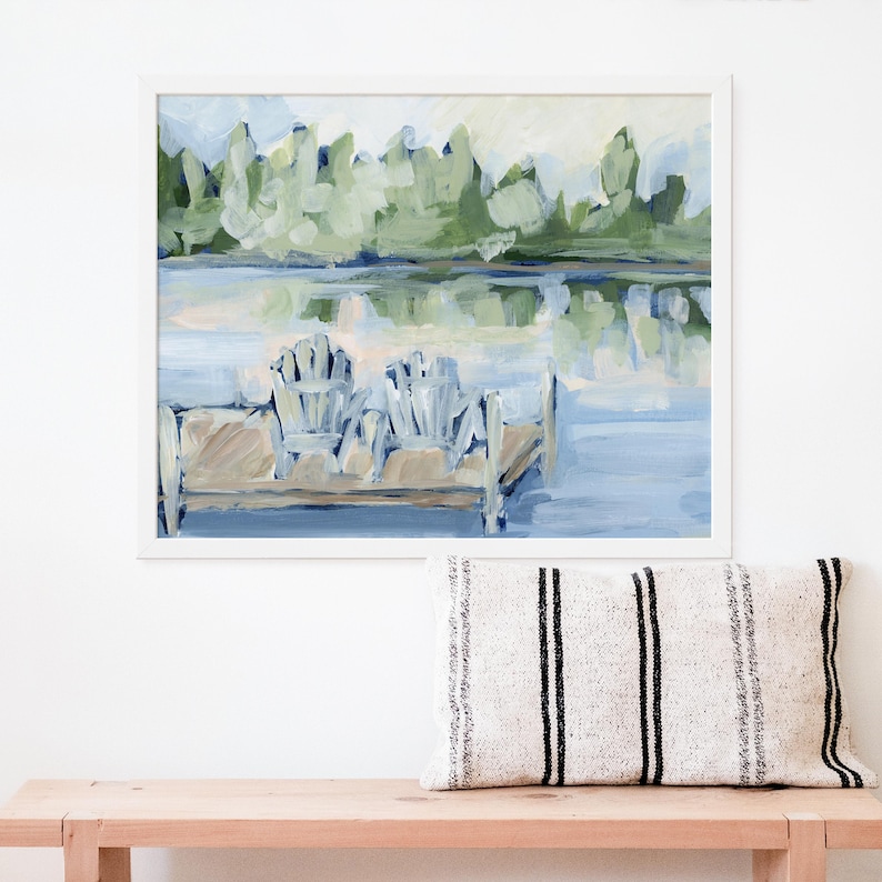 Pode incluir: Uma pintura de duas cadeiras Adirondack brancas em um cais de madeira com vista para um lago com &aacute;rvores verdes ao fundo.