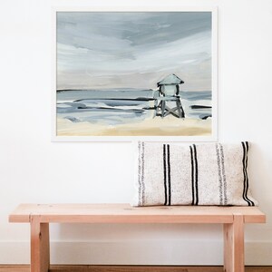May include: A framed painting of a beach scene with a lifeguard stand in the distance. The sky is a light blue with gray clouds, and the water is a light blue with white waves. The sand is a light yellow.