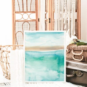 May include: A framed watercolor painting of a seascape with a light blue sky, brown sand, and turquoise water.