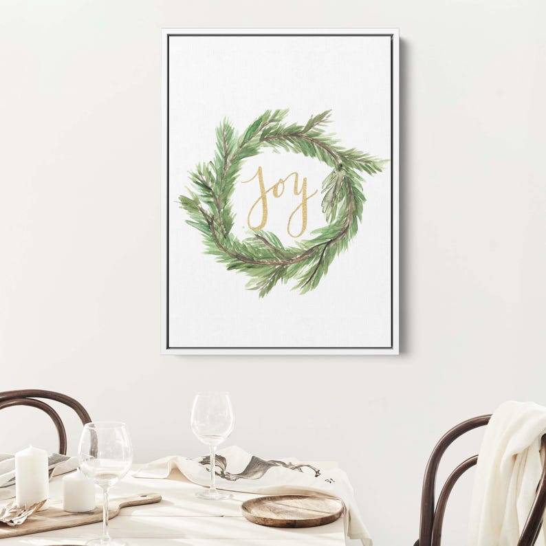 May include: A framed print featuring a watercolor wreath of green leaves. The word "Joy" is written in gold script within the wreath. The artwork is displayed on a white background, with a black frame.