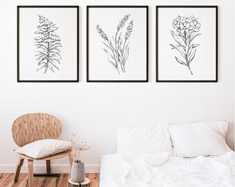 May include: Three black framed prints of botanical line drawings on a white wall. The prints feature a single flower or plant in each frame. The prints are hanging above a white bed with a wooden headboard and a white duvet.