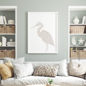 May include: A white framed print of a stylized line drawing of a heron in a light brown color. The print is hanging on a light green wall above a white couch with beige pillows.