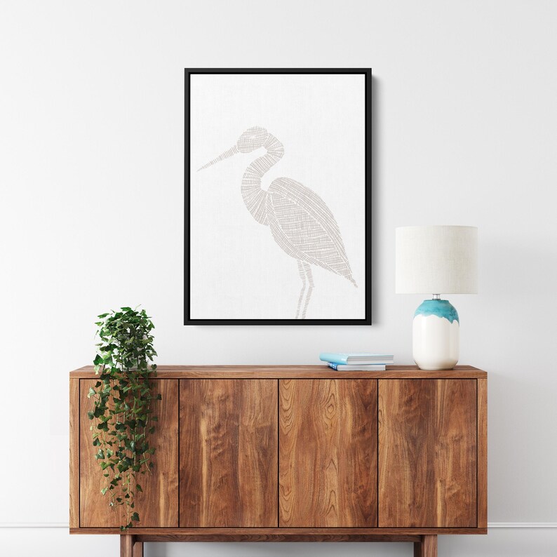 May include: A framed print of a white heron with a detailed line art design. The heron is standing on one leg with its head turned to the side. The print is hanging on a white wall above a wooden cabinet.