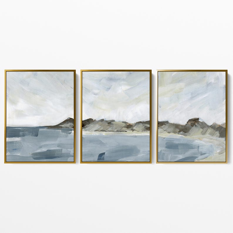 May include: Set of three framed abstract landscape paintings. Each piece features a muted color palette of blues, grays, and beige, depicting a seascape with mountains and sky. The gold frames add a touch of elegance to the artwork.