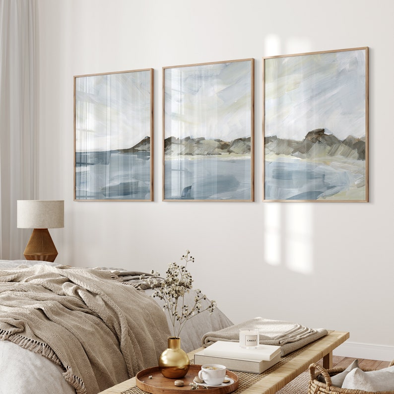 May include: Three framed abstract landscape paintings. Each features a serene coastal scene with muted blues, grays, and beige tones. The artwork is displayed in a bedroom setting, with a neutral color palette and natural wood frames.
