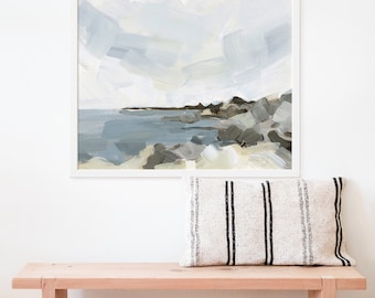 May include: A framed abstract painting of a seascape with a rocky shoreline and a cloudy sky. The painting is in shades of blue, gray, and white. The painting is hanging on a white wall above a wooden bench with a striped pillow.