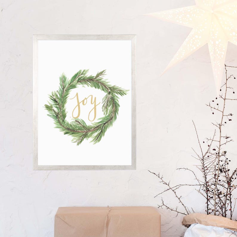 May include: A framed print featuring a watercolor wreath of green leaves. The word "joy" is written in gold script within the wreath. The frame is silver, and the background is white. The print is a decorative item.