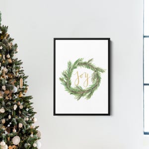 May include: A framed print featuring a watercolor wreath of green pine needles. The word "joy" is written in gold script within the wreath. The artwork is displayed on a white wall, with a Christmas tree visible on the left.