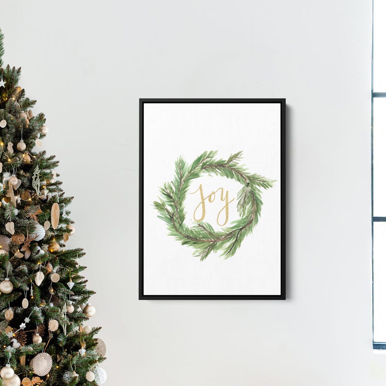 May include: A framed print featuring a watercolor wreath of green pine needles. The word "joy" is written in gold script within the wreath. The artwork is displayed on a white wall, with a Christmas tree visible on the left.