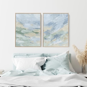 May include: Two abstract paintings in a light blue, green, and beige color scheme. The paintings are framed in a light wood frame and are hanging on a white wall above a bed with light blue bedding.