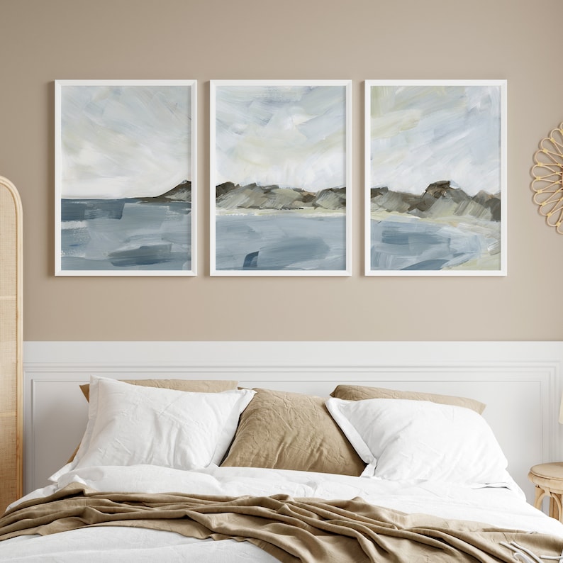 May include: Three framed abstract landscape paintings. Each features a coastal scene with blue water, gray mountains, and a light sky. The paintings are in white frames and hang above a bed with white and tan bedding.