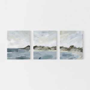 May include: Set of three abstract landscape paintings. Each canvas features a serene scene with a blue body of water, a brown and gray landmass, and a light blue and white sky. The artwork is displayed on a white background.
