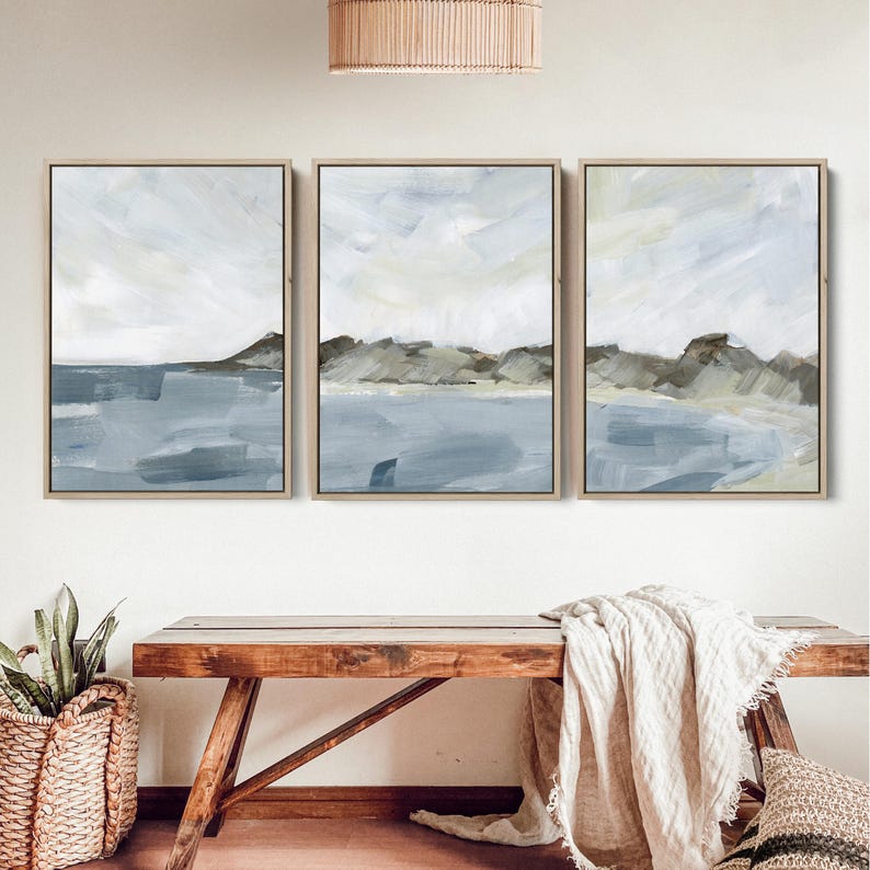 May include: Three framed abstract landscape paintings. Each features a coastal scene with blue water, gray-brown hills, and a light sky. The paintings are in light wood frames, creating a cohesive set for wall decor.