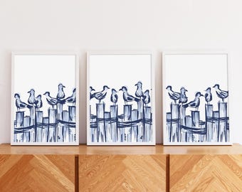 Puede incluir: Tres impresiones artísticas enmarcadas con una ilustración en acuarela de gaviotas posadas en un muelle azul. La obra de arte tiene un estilo minimalista con un fondo blanco y una combinación de colores azul y blanco. Las impresiones se exhiben en un mueble de madera.