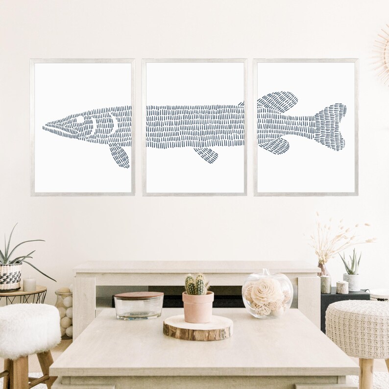 May include: A set of three framed prints featuring a stylized blue fish design. The fish is made up of a series of small lines, creating a textured effect. The prints are hung on a white wall above a fireplace.