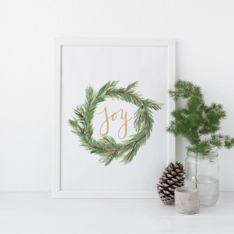 May include: A framed print featuring a watercolor wreath of green pine needles with the gold word "Joy" in the center. A pine cone, a small concrete candle holder, and a glass jar with greenery are next to the frame.