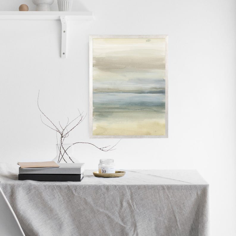 May include: A framed abstract watercolor painting with shades of blue, green, and brown. The painting depicts a landscape with a horizon line and a sky with clouds. The frame is silver and has a white mat.