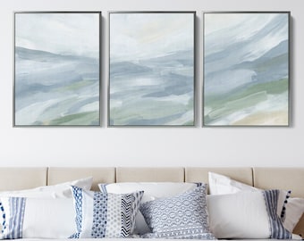 May include: A set of three abstract landscape paintings in shades of blue and green. The paintings are framed in silver and hung on a white wall above a beige headboard.