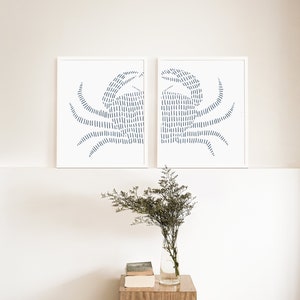 May include: Two white framed prints of blue crab illustrations. The crabs are made of thin lines and are facing each other. The prints are hanging on a white wall above a wooden table with a vase of flowers.