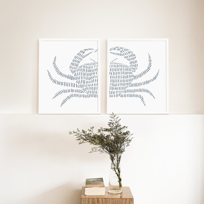 May include: Two white framed prints of blue crab illustrations. The crabs are made of thin lines and are facing each other. The prints are hanging on a white wall above a wooden table with a vase of flowers.