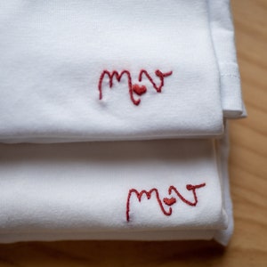 May include: Two white t-shirts folded on top of each other. The top shirt has the word "mom" embroidered in red with a small heart in the middle of the "o". The bottom shirt has the same embroidery.