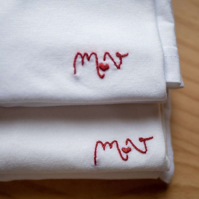 May include: Two white t-shirts folded on top of each other. The top shirt has the word "mom" embroidered in red with a small heart in the middle of the "o". The bottom shirt has the same embroidery.