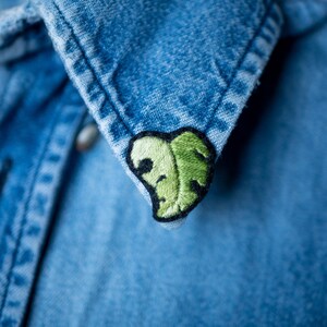 Peut inclure: Un gros plan sur un col de chemise en jean avec un patch brodé de feuille verte.