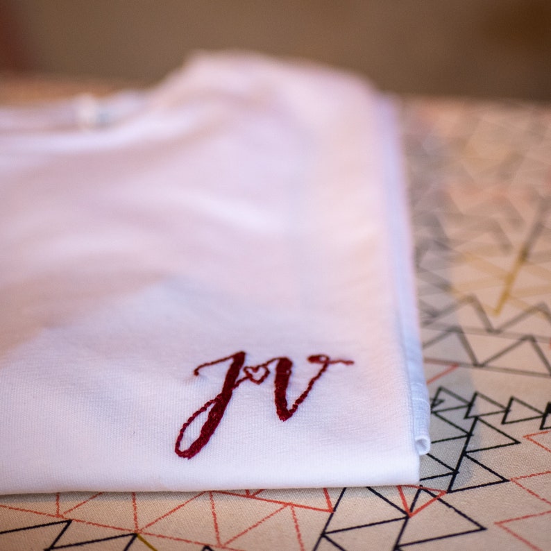 May include: White t-shirt with a red embroidered "JV" monogram with a small heart in the middle of the letters.