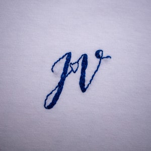 May include: Embroidered "Jr" with a small heart in the center of the "J". The embroidery is done in blue thread on a white fabric.