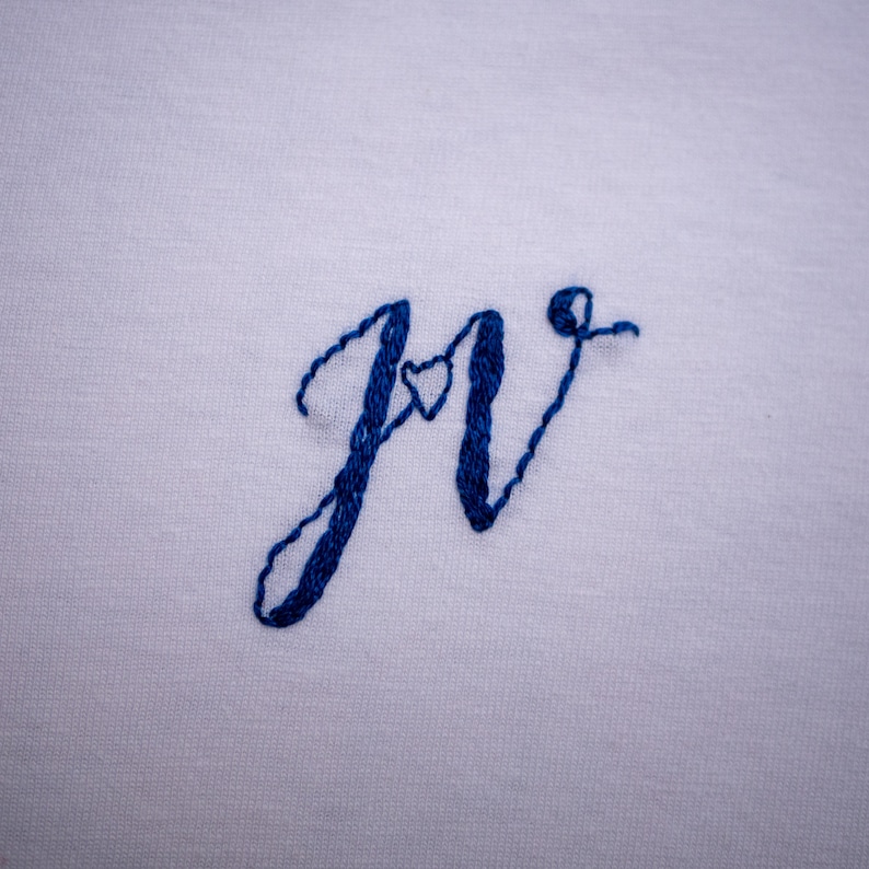 May include: Embroidered "Jr" with a small heart in the center of the "J". The embroidery is done in blue thread on a white fabric.