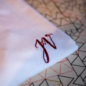 May include: White fabric with a red embroidered monogram "JG" in a cursive script.