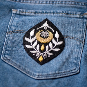 May include: A black denim pocket with a gold and white embroidered patch featuring a crescent moon, eye, and floral design.