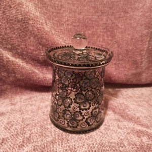 May include: A clear glass jar with a lid and a small spoon. The jar is decorated with a black and green floral pattern.