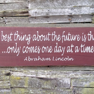 May include: A red wooden sign with white lettering that reads "The best thing about the future is that it... only comes one day at a time. Abraham Lincoln".