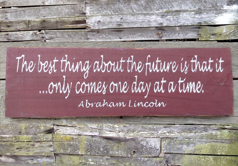May include: A red wooden sign with white lettering that reads "The best thing about the future is that it... only comes one day at a time. Abraham Lincoln".