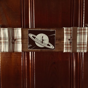 May include: A wooden wall-mounted coat rack with two plaid fabric straps and leather straps. The rack has two hooks on each side and a black and white image of a planet with rings in the center.