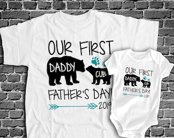 May include: White t-shirt and onesie with black bear silhouettes and the text "Our First Daddy/Cub Father's Day 2019".
