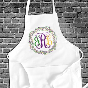 May include: White kitchen apron with a pocket and adjustable straps. The apron features a purple, green, and gold embroidered monogram in the shape of a wreath with fleur-de-lis accents.