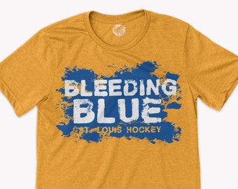 May include: A mustard yellow t-shirt with a blue and white graphic print that says "Bleeding Blue" and "St. Louis Hockey".