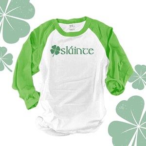 May include: White and green baseball style t-shirt with a four-leaf clover and the word "sláinte" printed on the front.