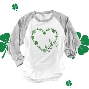 May include: A white and gray baseball tee shirt with a green shamrock heart design and the word "lucky" written inside the heart.