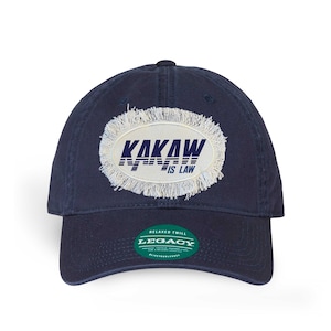 May include: A navy blue baseball cap with a distressed white oval patch that reads "KAKAW IS LAW" in navy blue lettering. The cap has a green label that reads "RELAXED TWILL LEGACY".