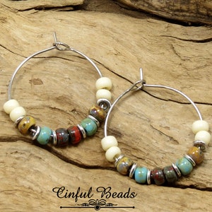 May include: A pair of silver hoop earrings with a mix of white, brown, blue, and red beads. The earrings are on a wooden surface.