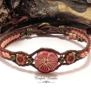 May include: A handcrafted bracelet featuring a central pink flower bead with a detailed design, flanked by smaller flower beads. The bracelet has a brown leather band with peach-colored beads and antique brass accents. The text "Cinful Beads" is visible.