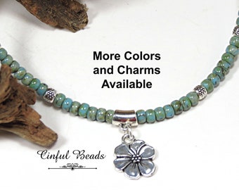 May include: A turquoise beaded necklace with a silver flower pendant. The necklace features small, round beads and silver accents. The text "More Colors and Charms Available" is visible.