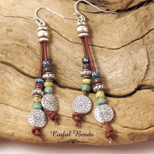 May include: A pair of long, dangle earrings with brown leather cords, silver beads, and colorful ceramic beads. The earrings feature three silver circular charms with an intricate floral design.
