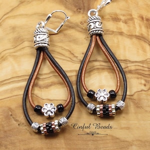 May include: A pair of dangle earrings with brown and black leather cords, silver beads, and silver flower charms. The earrings are accented with black beads and a silver cylinder bead with black and copper accents. The earrings are labeled "Cinflul Beads".