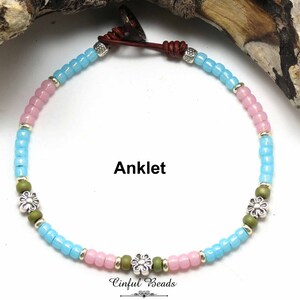 May include: A light blue, pink, and green beaded anklet with silver flower charms. The anklet is tied with a brown leather cord and has the text "Anklet" printed below it.